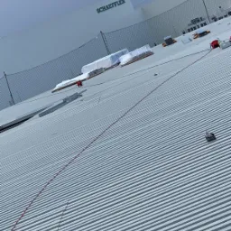 Falisty dach przemysłowy z elementami instalacji odgromowej w trakcie montażu, widoczne zabezpieczenia siatkowe i materiały budowlane na dachu. W tle budynek z logo Schaeffler.