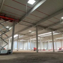 Wnętrze hali magazynowej w trakcie budowy z widoczną czerwoną platformą podnośnikową Mateco z pracownikami przy suficie oraz licznymi słupami nośnymi i instalacjami pod stropem.