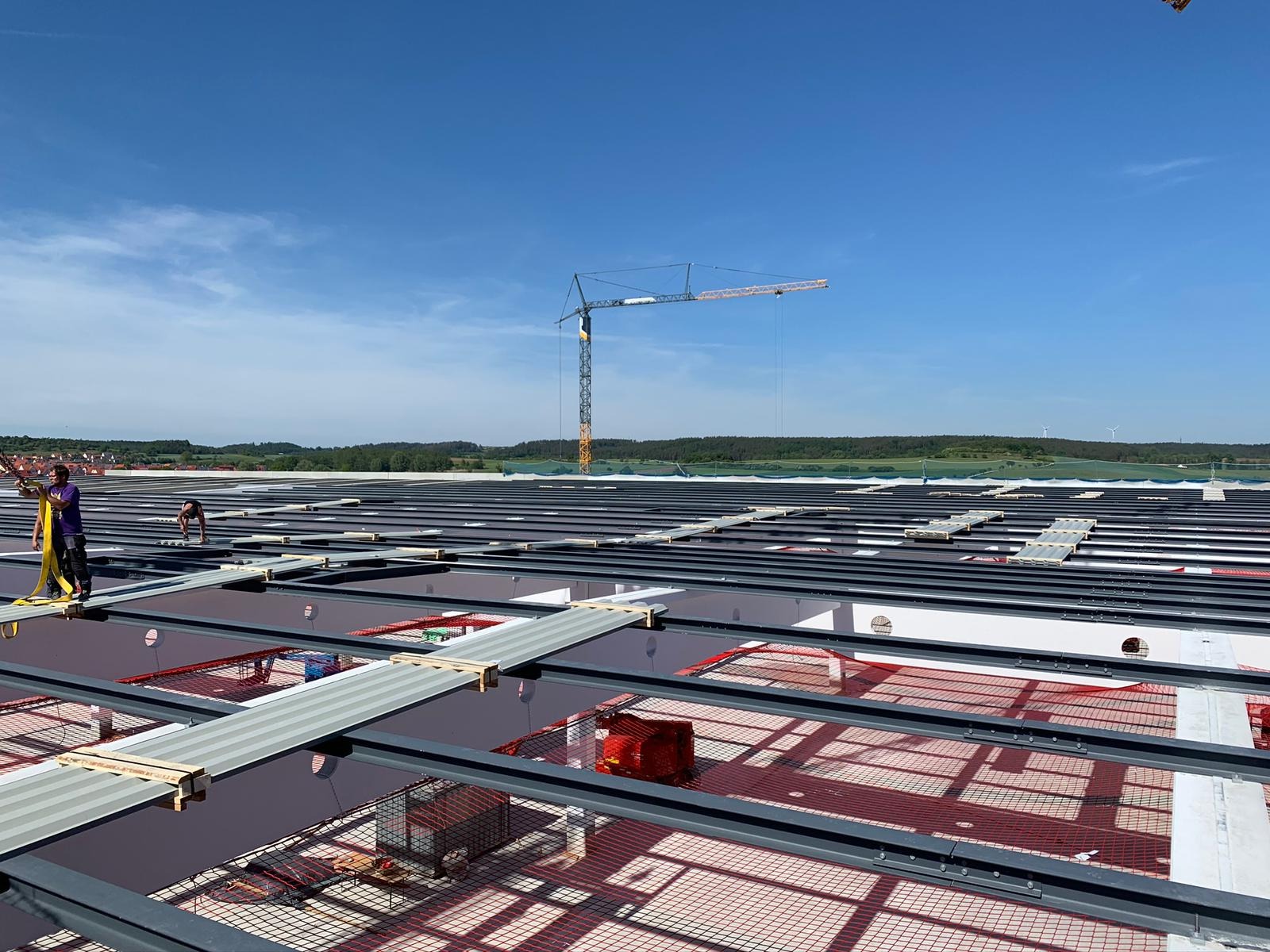 Montaż konstrukcji wsporczej pod panele fotowoltaiczne na dachu hali przemysłowej, widoczni pracownicy w uprzężach, dźwig w tle, słoneczny dzień.