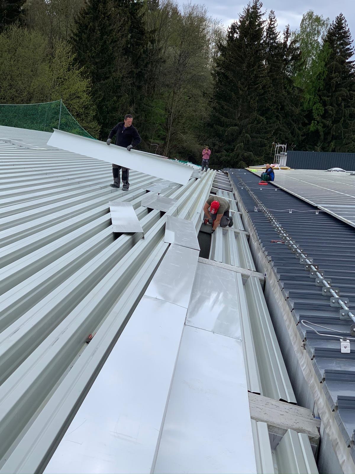 Montaż paneli na dachu: dekarz w czerwonym kasku mocuje blachę trapezową wkrętarką akumulatorową, drugi pracownik przenosi element dachu, w tle drzewa i częściowo zainstalowane panele słoneczne.