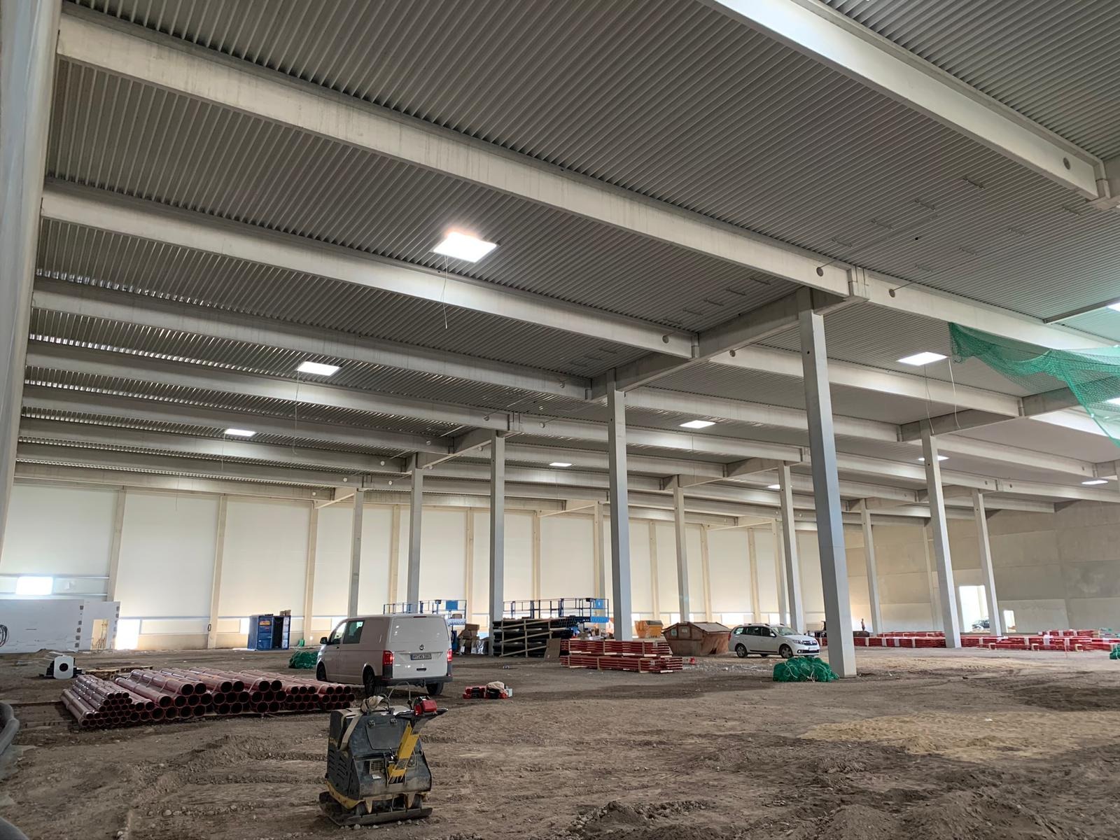 Wnętrze hali produkcyjnej w trakcie montażu oświetlenia, widoczne stalowe słupy, strop z blachy trapezowej, rzędy lamp oraz przygotowane rury i zagęszczarka wibracyjna na ziemi.