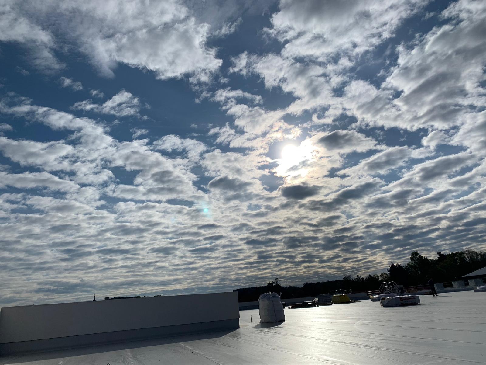 Widok na niebo z chmurami z poziomu płaskiego, białego dachu budynku. Na dachu widoczne elementy konstrukcyjne i materiały budowlane.