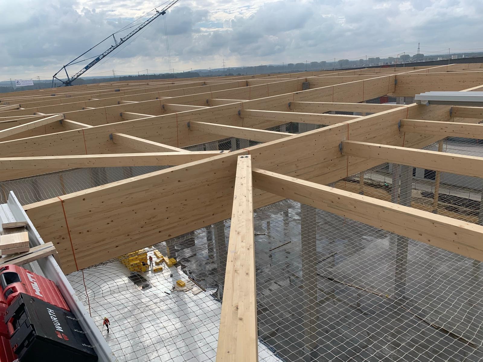 Widok z góry na konstrukcję drewnianą dachu hali przemysłowej, z widocznymi narzędziami Wurth i Hilti na pierwszym planie, zabezpieczona siatką, w tle żuraw budowlany i pochmurne niebo.