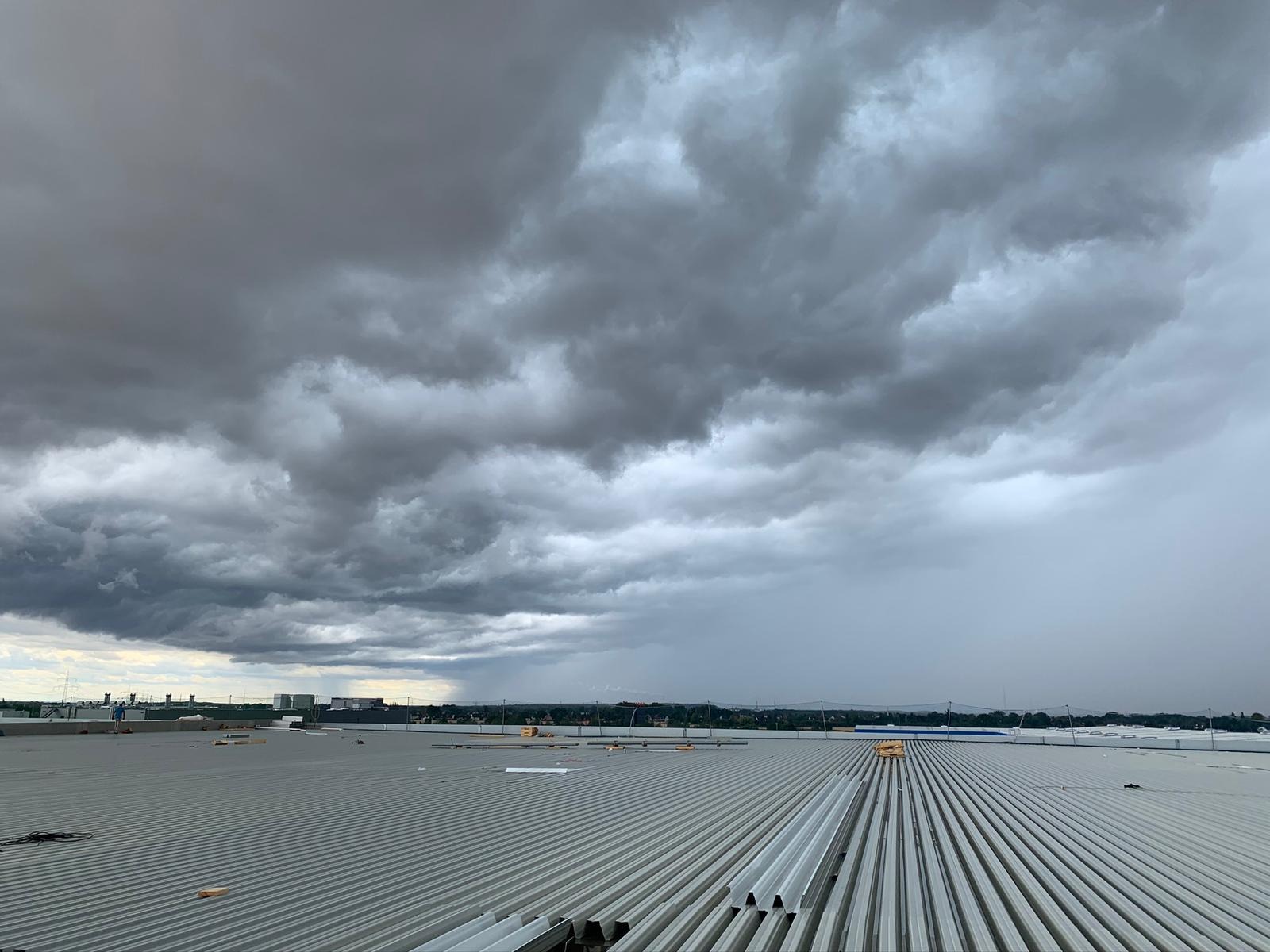 Szara, burzowa chmura nad falistym, metalowym dachem budynku przemysłowego z widocznymi elementami instalacji odgromowej i fragmentem krajobrazu w tle.