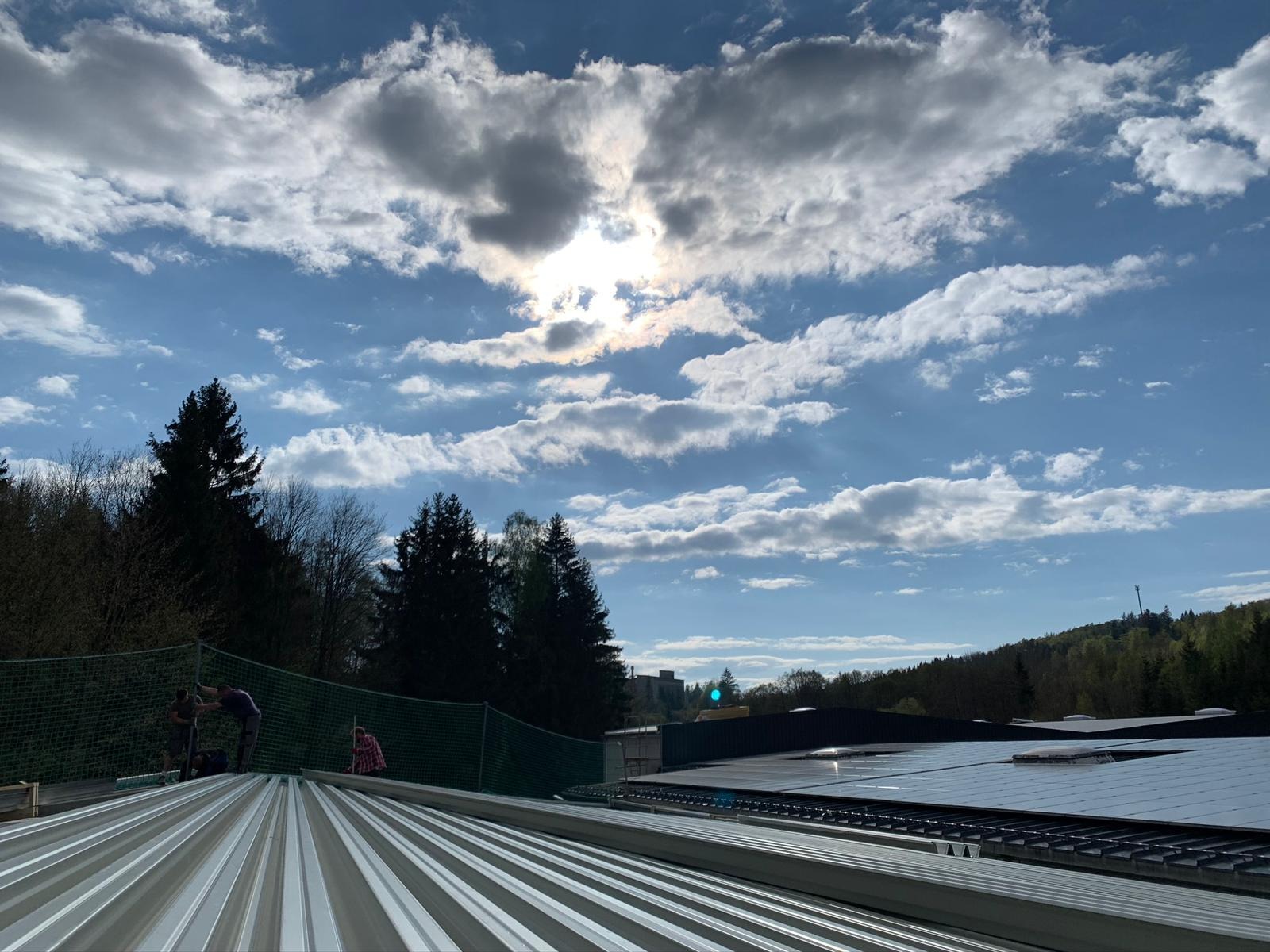 Montaż paneli fotowoltaicznych na blaszanym dachu z widokiem na las i błękitne niebo z chmurami, zabezpieczony siatką ochronną.