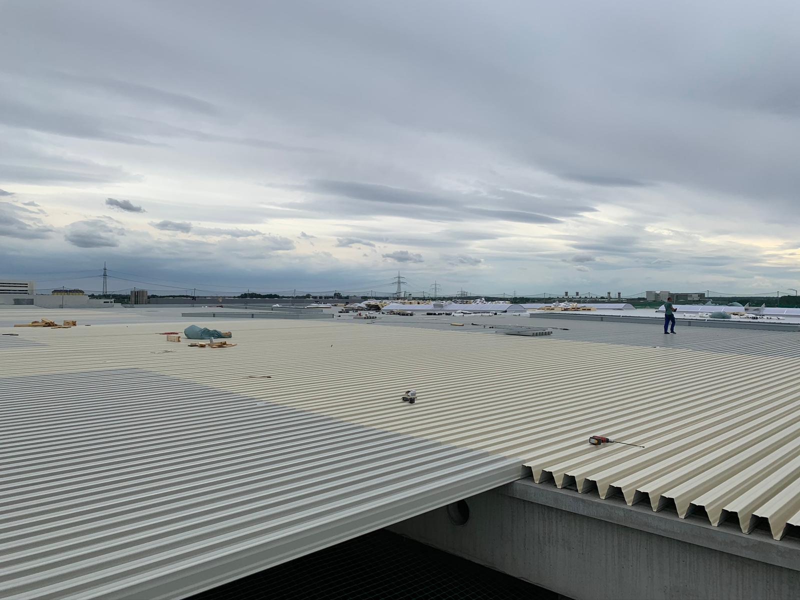 Szeroki kadr na falisty dach przemysłowy z widocznym pracownikiem w oddali oraz narzędziem na dachu, na tle pochmurnego nieba i elementów infrastruktury.