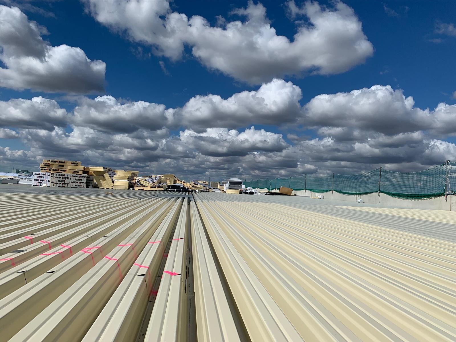 Montaż paneli fotowoltaicznych na płaskim dachu z widoczną konstrukcją wsporczą, materiałami izolacyjnymi i siatką zabezpieczającą, pod błękitnym niebem z chmurami.