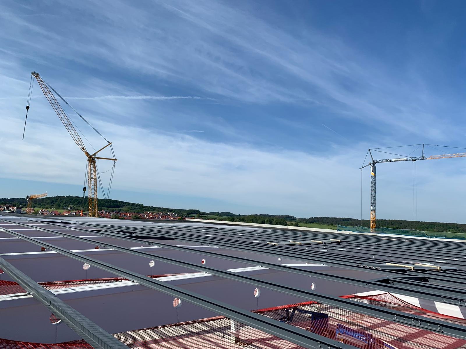 Montaż paneli fotowoltaicznych na dachu hali przemysłowej, widoczne stalowe konstrukcje montażowe, siatka zabezpieczająca i dwa żurawie budowlane na tle błękitnego nieba z chmurami.