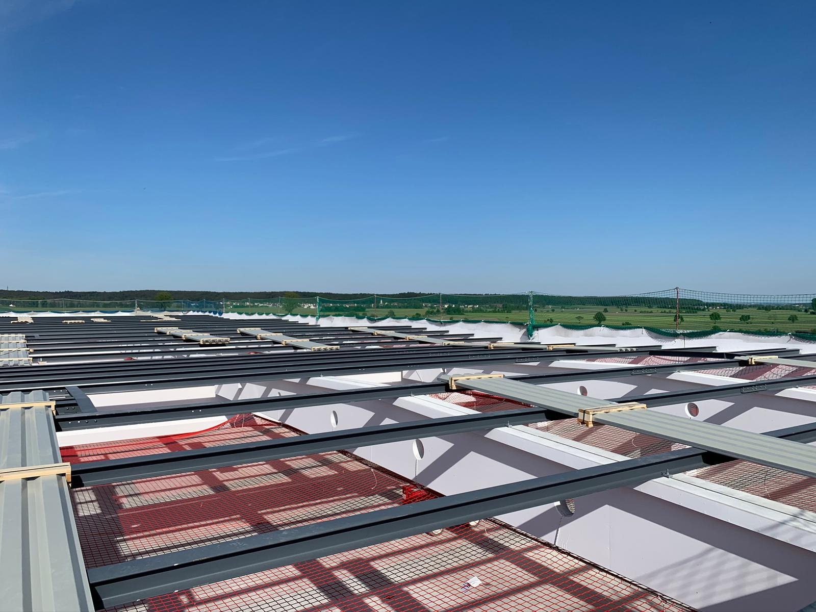 Widok z góry na konstrukcję dachu z metalowymi belkami, siatką zabezpieczającą i elementami izolacyjnymi w trakcie montażu, na tle błękitnego nieba i zielonego krajobrazu.