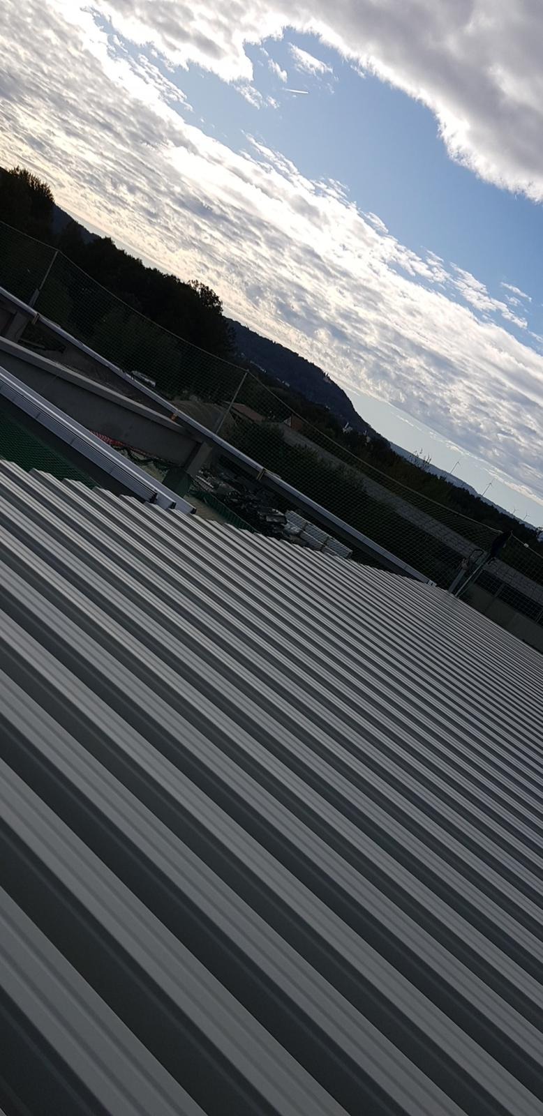 Szare blaszane pokrycie dachu z widocznymi mocowaniami, na tle błękitnego nieba z chmurami i zalesionych wzgórz w oddali.