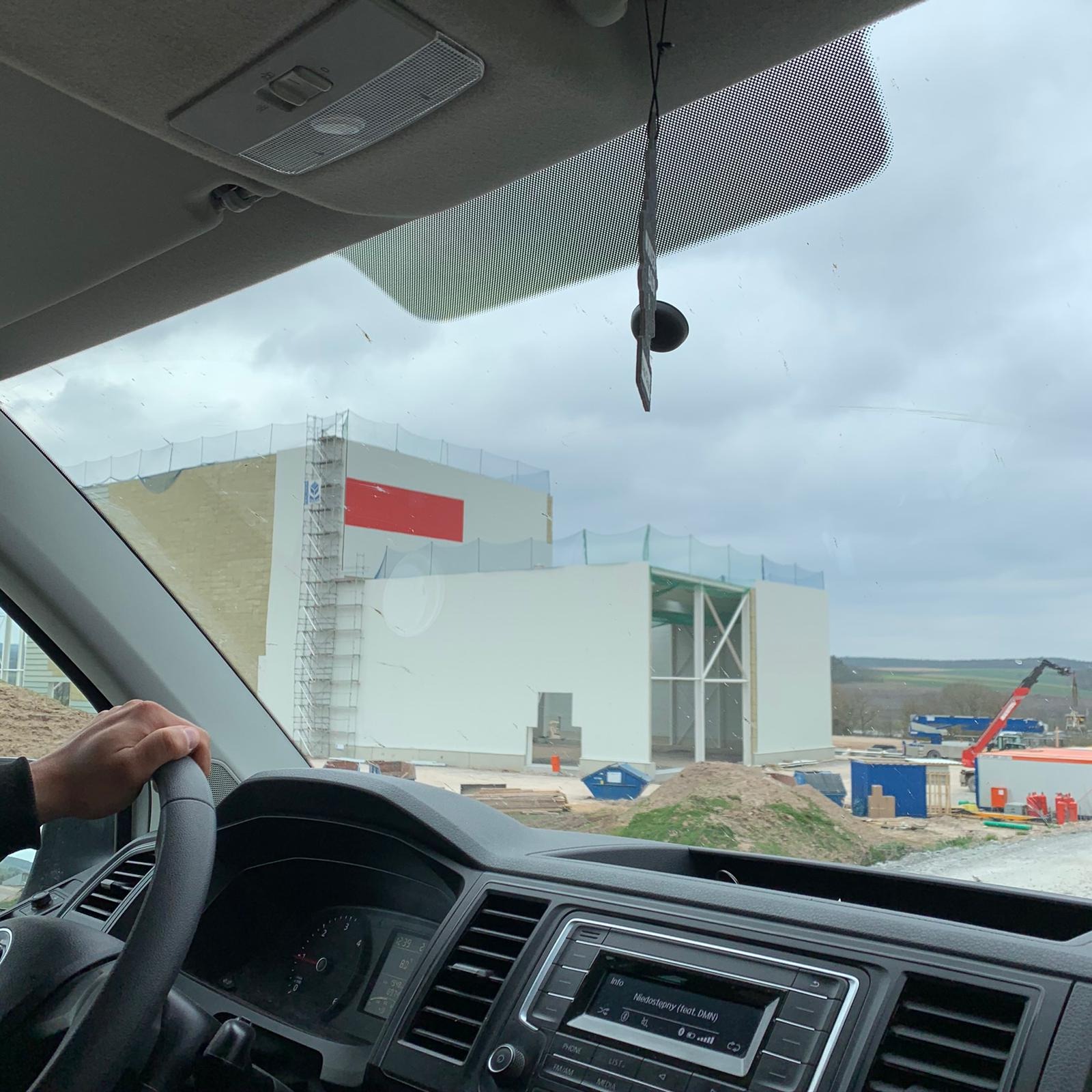 Widok z wnętrza samochodu na budynek w trakcie budowy, widoczny fragment rusztowania i czerwony panel na fasadzie, teren budowy z kontenerami i żurawiem.