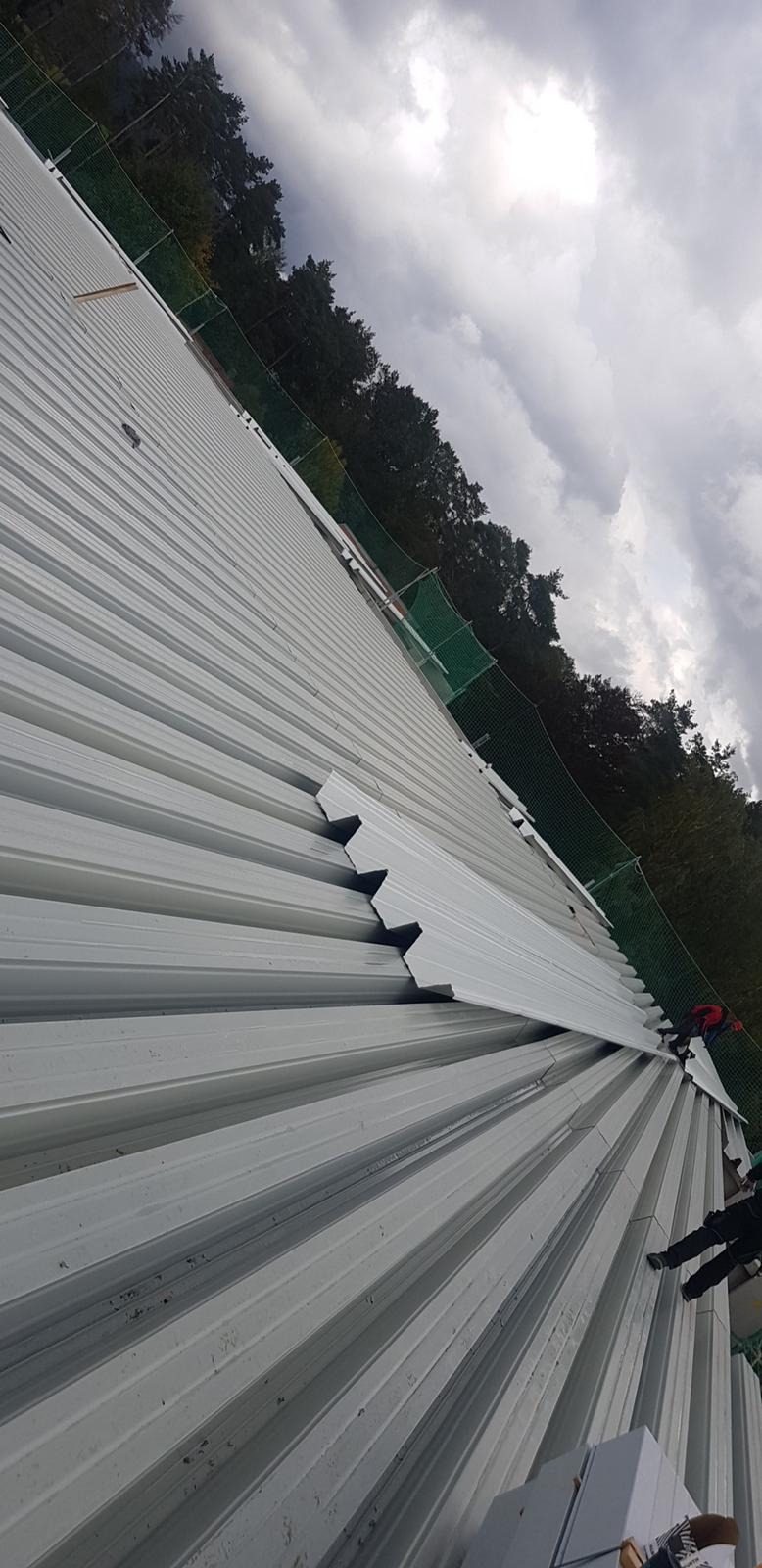 Montaż białych paneli blaszanych na dachu budynku, widoczne fragmenty lasu i pochmurne niebo w tle.