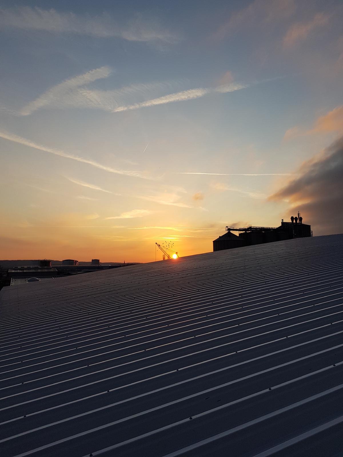 Metalowy dach przemysłowy z widocznym zachodem słońca i konstrukcją na horyzoncie, smugi kondensacyjne na niebie.