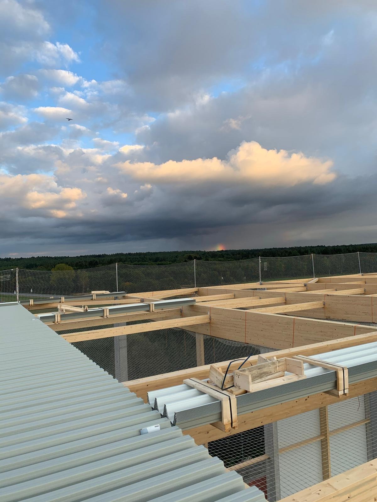 Konstrukcja dachu z drewnianych belek i metalowych profili, zabezpieczona siatką, z widocznym fragmentem tęczy na horyzoncie pod burzowymi chmurami.