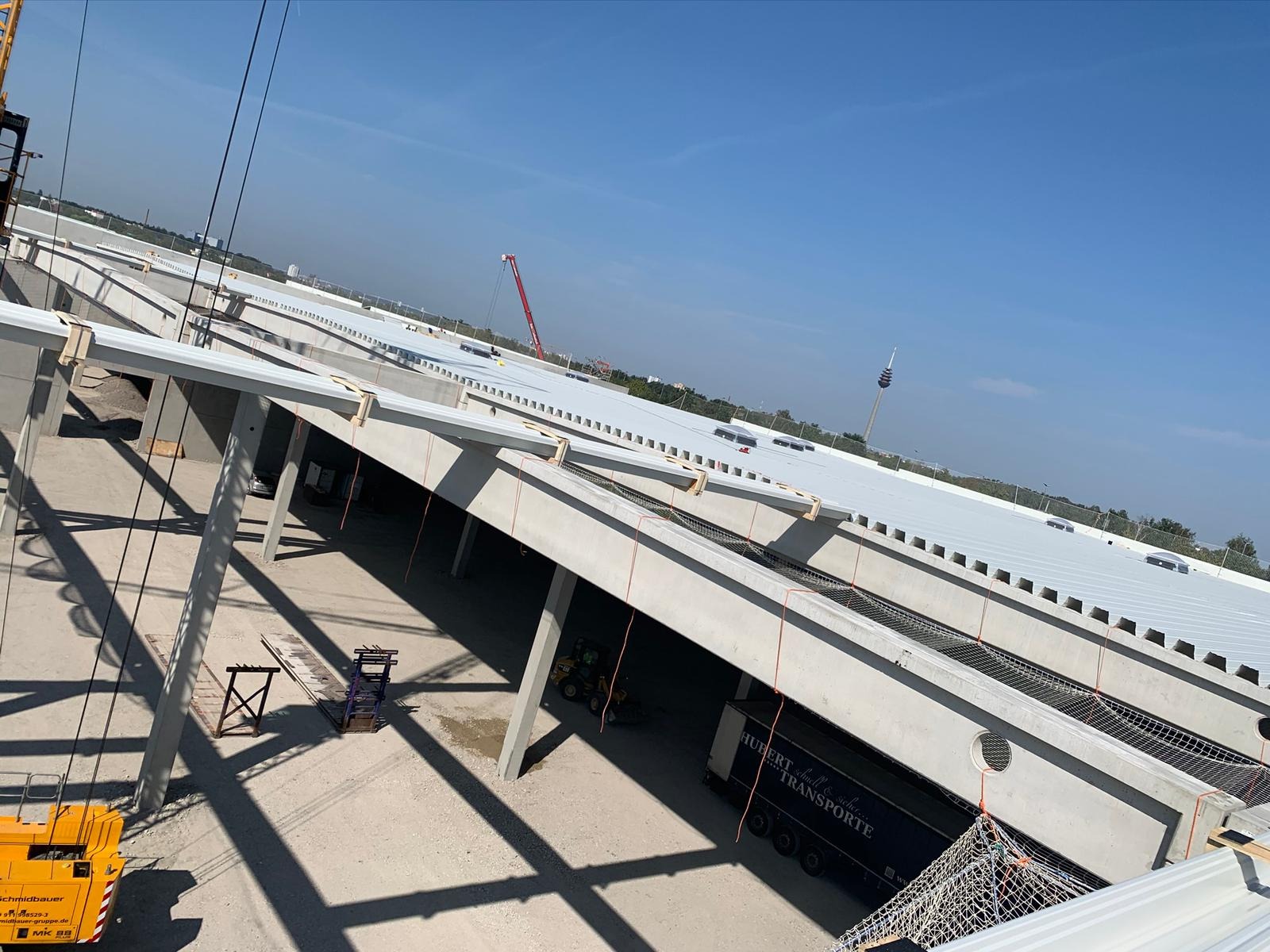 Budowa hali przemysłowej z widocznymi betonowymi konstrukcjami, częściowo zamontowanym dachem z blachy trapezowej, dźwigiem budowlanym i ciężarówką transportową na placu budowy w słoneczny dzień.