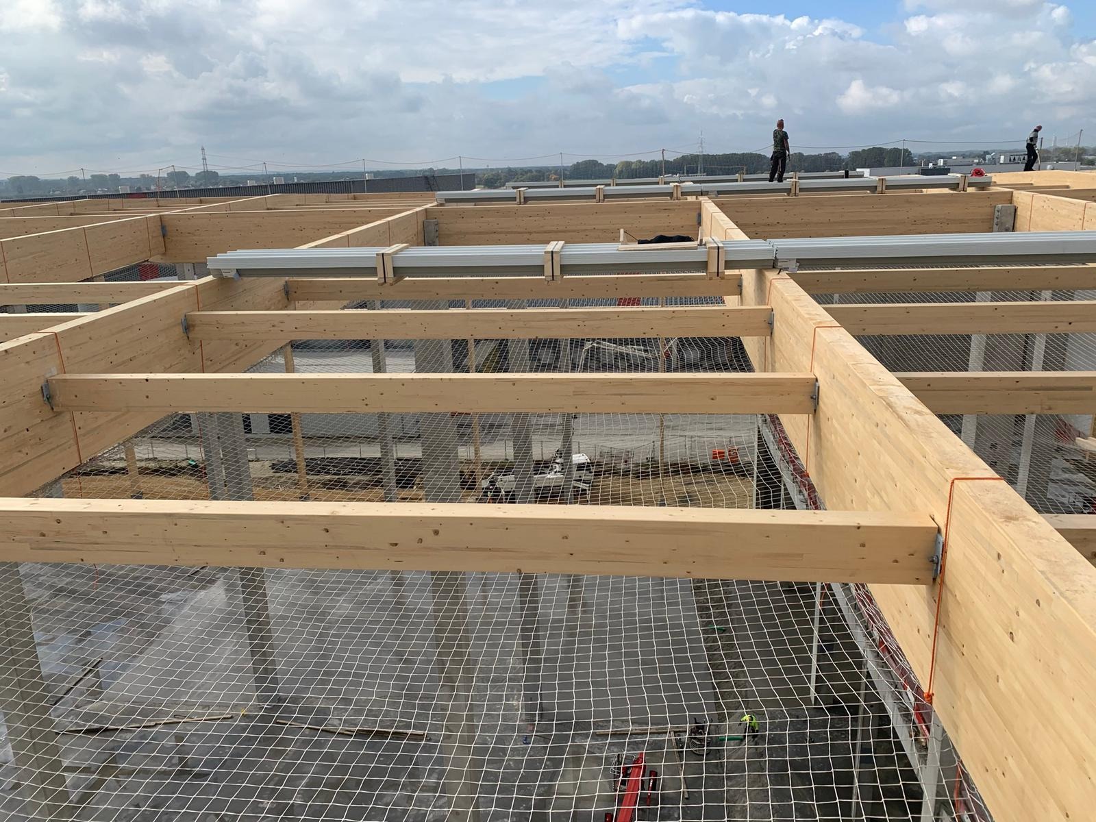 Konstrukcja drewniana dachu budynku w trakcie montażu, zabezpieczona siatką ochronną, z widocznymi pracownikami na górze i elementami instalacji.