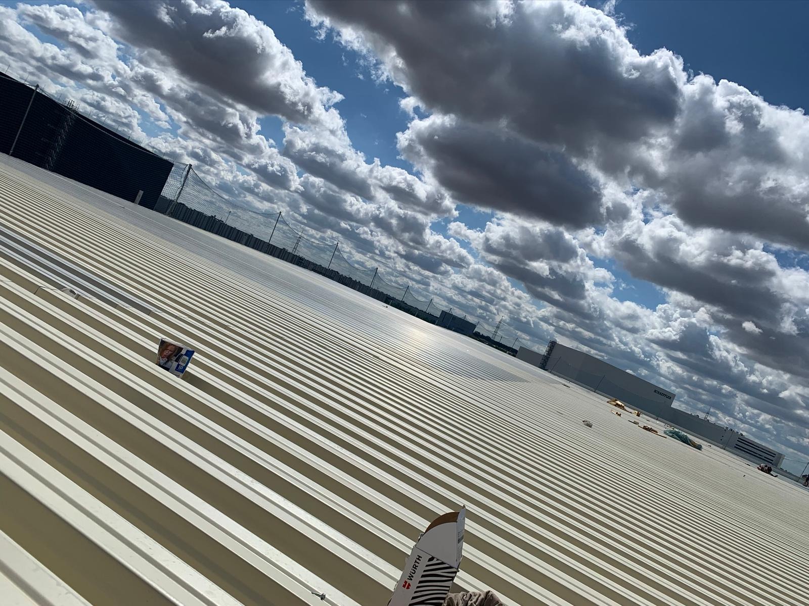 Jasna, blaszana konstrukcja dachu przemysłowego z widocznymi elementami mocującymi, na tle błękitnego nieba z chmurami, z elementami opakowania Wurth na pierwszym planie.