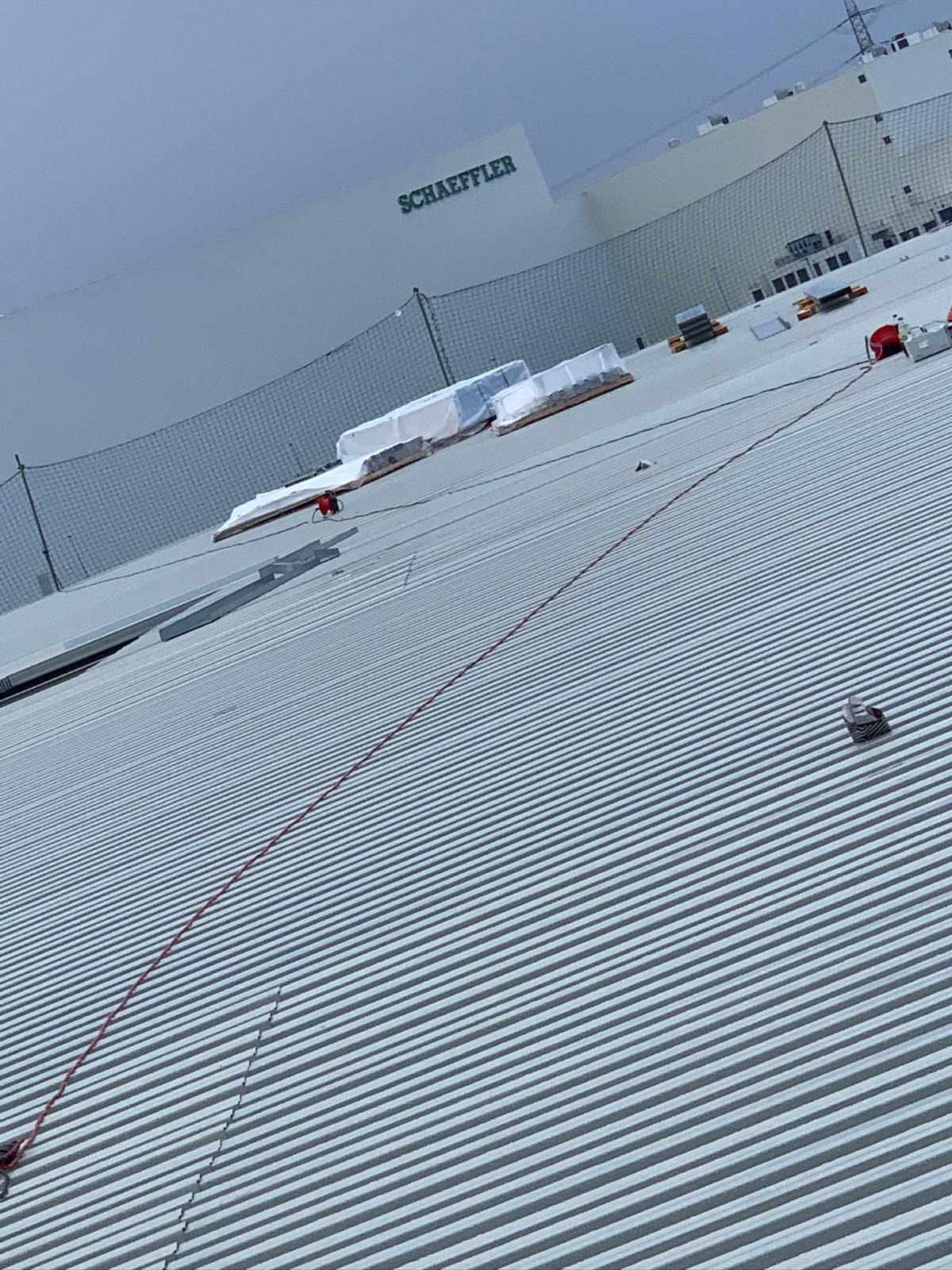 Falisty dach przemysłowy z elementami instalacji odgromowej w trakcie montażu, widoczne zabezpieczenia siatkowe i materiały budowlane na dachu. W tle budynek z logo Schaeffler.