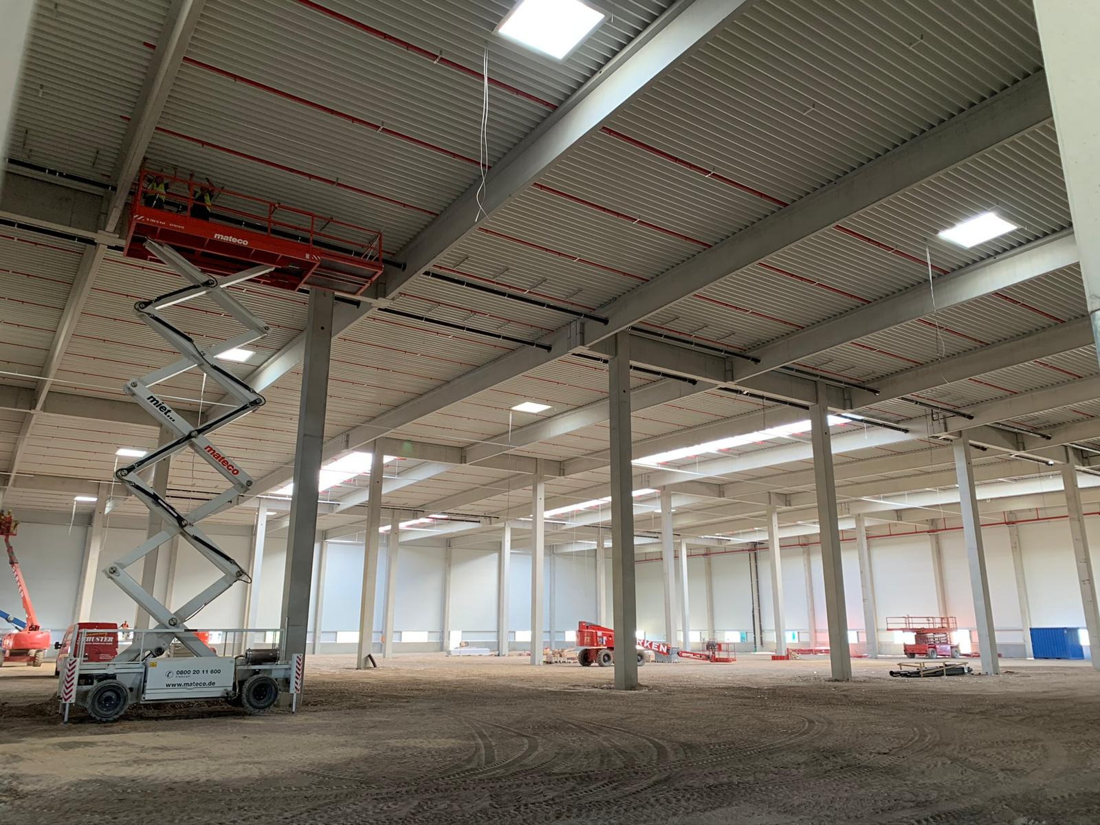 Wnętrze hali magazynowej w trakcie budowy z widoczną czerwoną platformą podnośnikową Mateco z pracownikami przy suficie oraz licznymi słupami nośnymi i instalacjami pod stropem.
