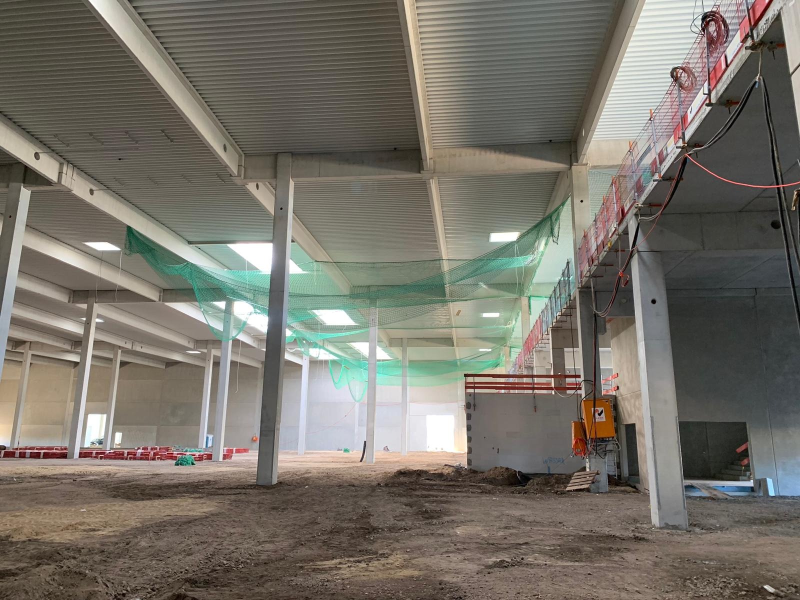 Wnętrze hali przemysłowej w trakcie budowy, widoczne stalowe konstrukcje stropu, słupy, siatka zabezpieczająca pod sufitem i prowizoryczne instalacje elektryczne na ścianach.