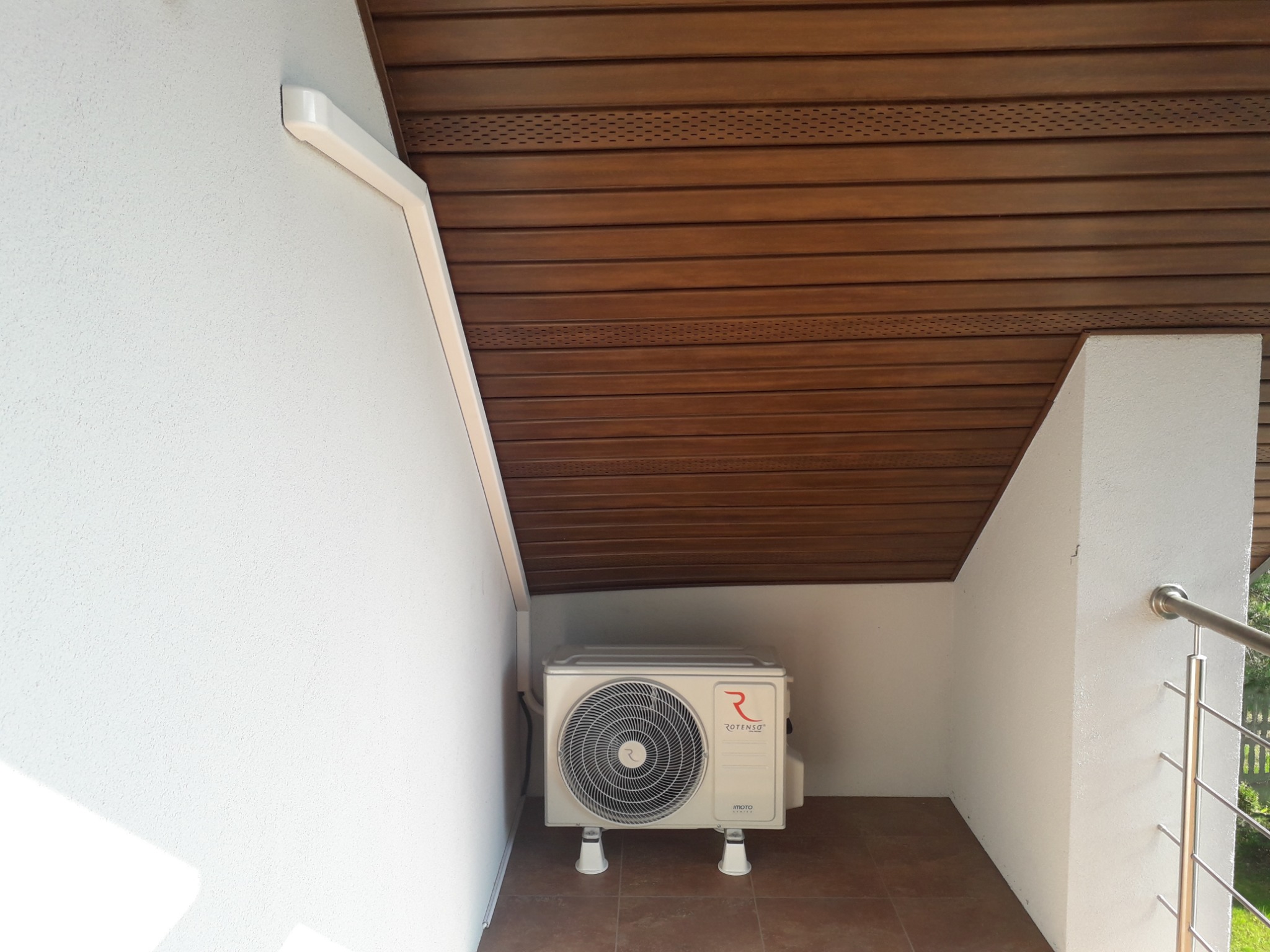 Zewnętrzna jednostka klimatyzacji Rotenso umieszczona w narożniku balkonu z drewnianym sufitem i terakotą na podłodze.