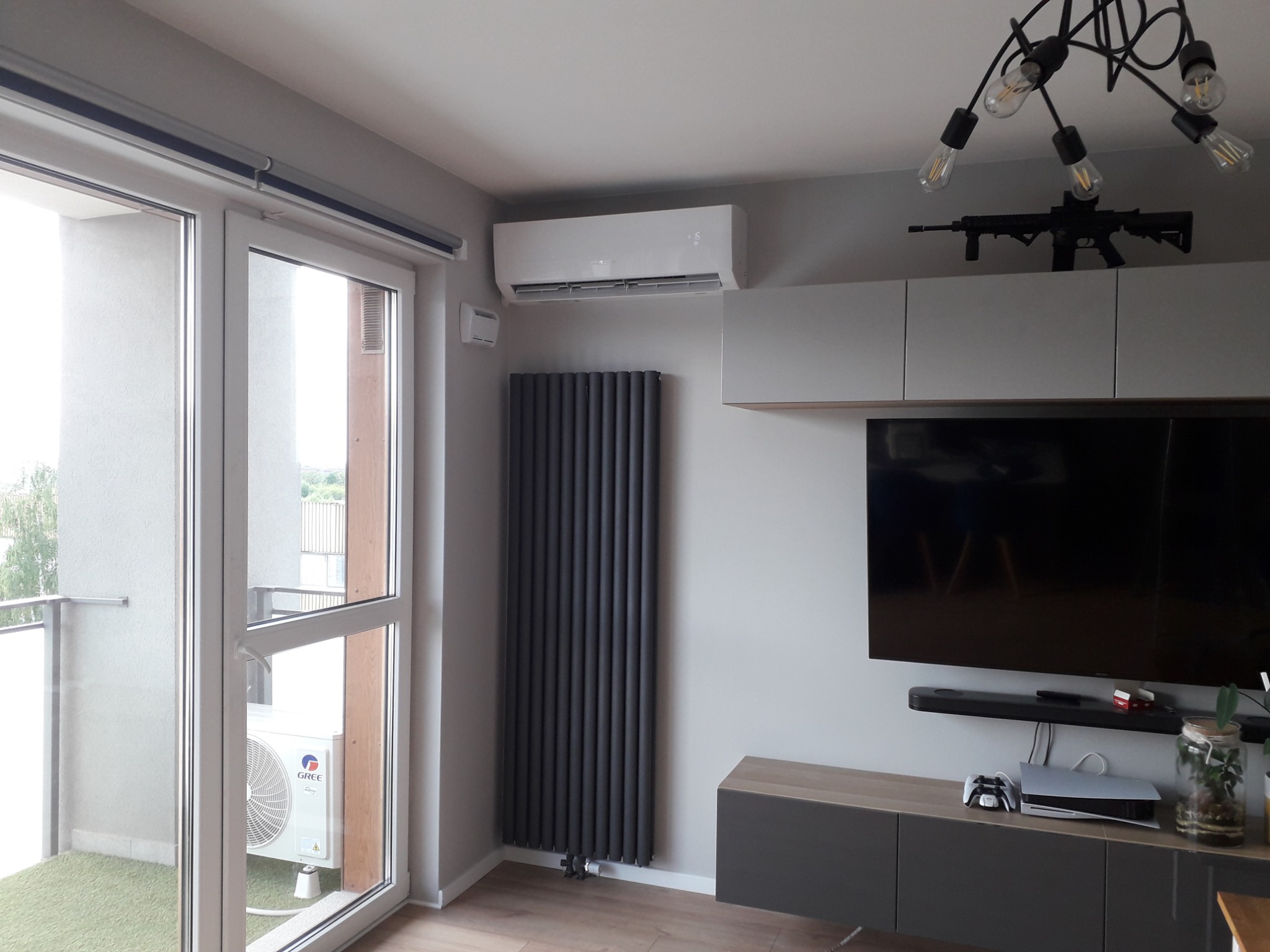 Nowoczesne wnętrze mieszkania z zamontowanym klimatyzatorem ściennym, grzejnikiem panelowym i widokiem na balkon z jednostką zewnętrzną klimatyzacji.