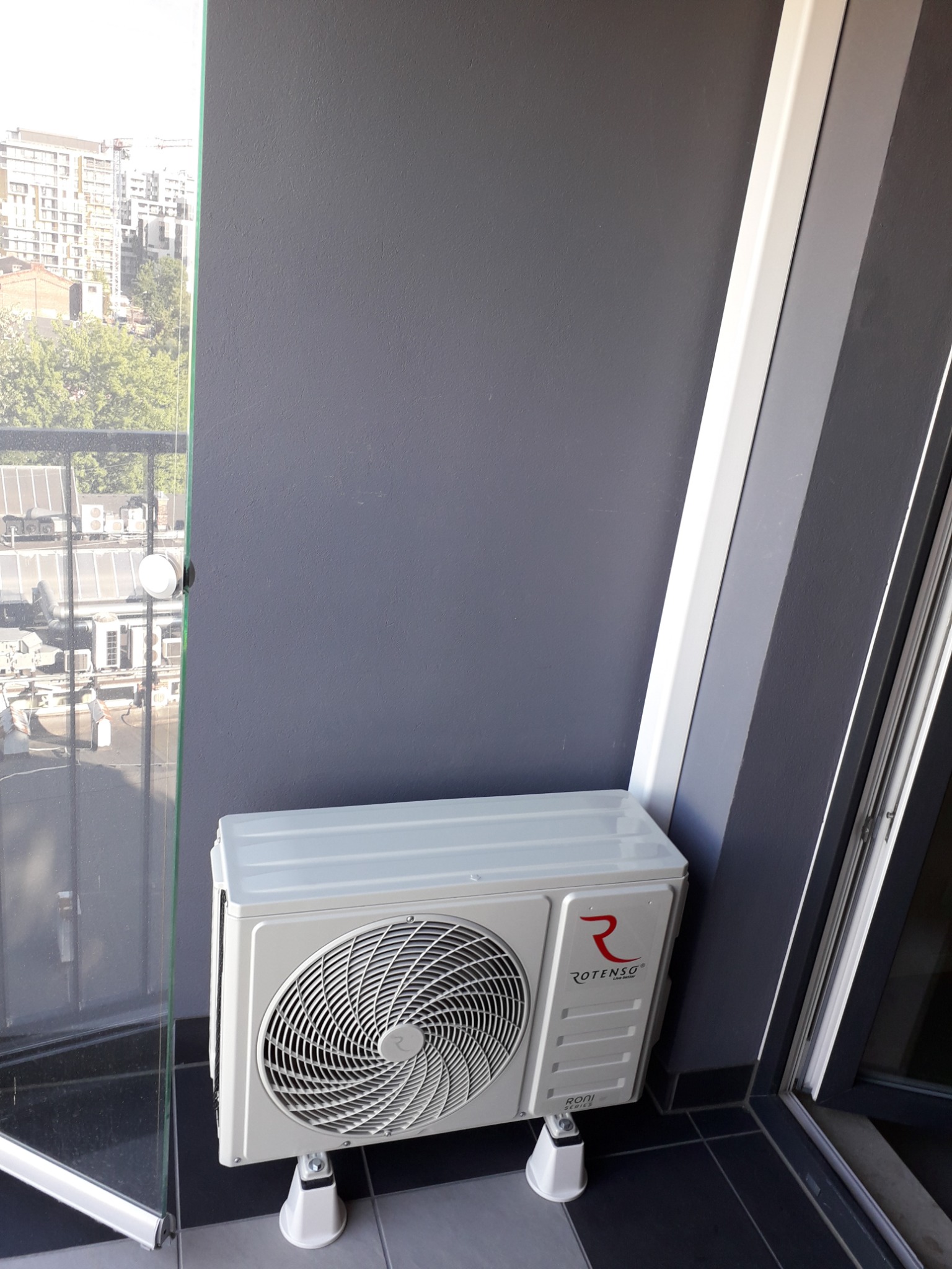 Biała jednostka zewnętrzna klimatyzatora Rotenso, model Roni Series, zamontowana na balkonie z widokiem na zabudowę miejską przez szklaną balustradę.