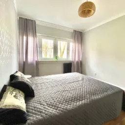 Jasna sypialnia z szarą pościelą w kratę, wzorzysta tapeta, okno z widokiem na drzewa i rattanowa lampa wisząca.