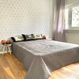 Jasna sypialnia z szarą pościelą w geometryczne wzory, poduszkami z motywem słoni, drewnianą podłogą i stolikiem nocnym z dekoracyjnymi kulami.