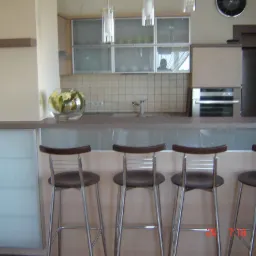 Nowoczesna aranżacja kuchni z barkiem, czterema wysokimi krzesłami barowymi ze stalowymi nogami i ciemnymi siedziskami, przeszklonymi szafkami wiszącymi oraz blatem z dekoracyjną misą z kwiatami.