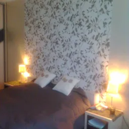 Sypialnia z łóżkiem, białymi poduszkami z motywem kwiatowym i tapetą w liście. Dwie lampki nocne ze światłem o ciepłej barwie oświetlają pomieszczenie.