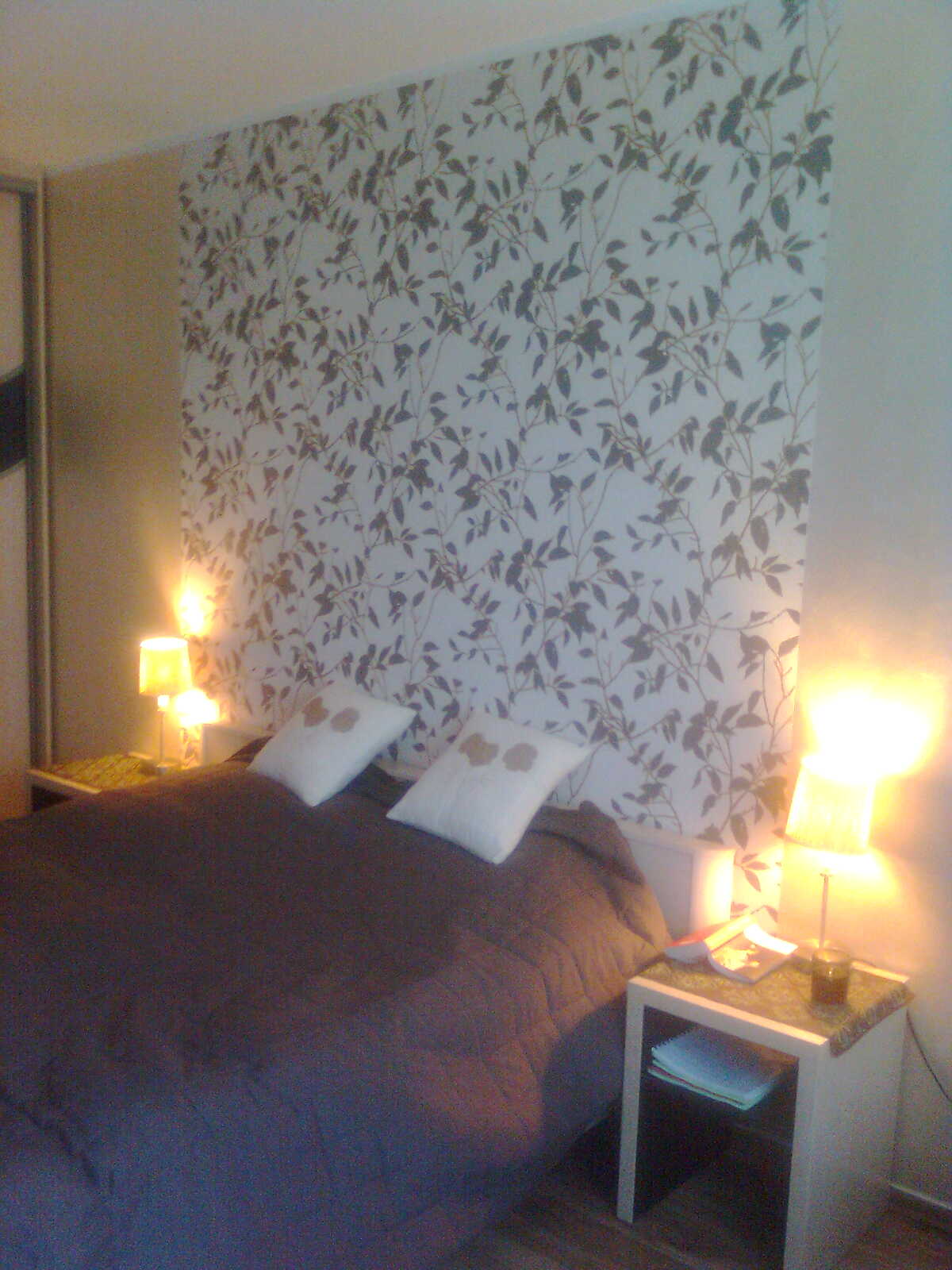 Sypialnia z łóżkiem, białymi poduszkami z motywem kwiatowym i tapetą w liście. Dwie lampki nocne ze światłem o ciepłej barwie oświetlają pomieszczenie.