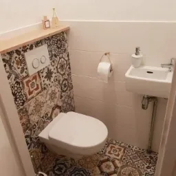 Mała łazienka z mozaikową glazurą na podłodze i ścianie za toaletą, biała umywalka narożna i półka z dekoracyjnymi flakonami.