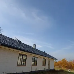 Dom jednorodzinny w trakcie budowy, z widocznym nowym dachem z czarnej dachówki ceramicznej i kominem, na tle błękitnego nieba i jesiennych drzew.