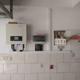 Instalacja elektryczna z inwerterem SOFAR, rozdzielnicą PV i otwartą skrzynką elektryczną; widoczne dłonie elektryka podłączającego przewody.