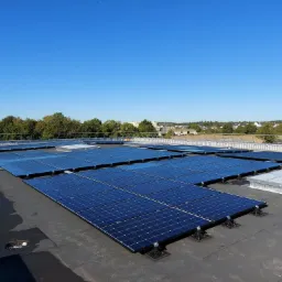 Rozległa instalacja paneli słonecznych na płaskim dachu z widoczną rolką kabla i okoliczną zabudową w tle, wykonana w słoneczny dzień.