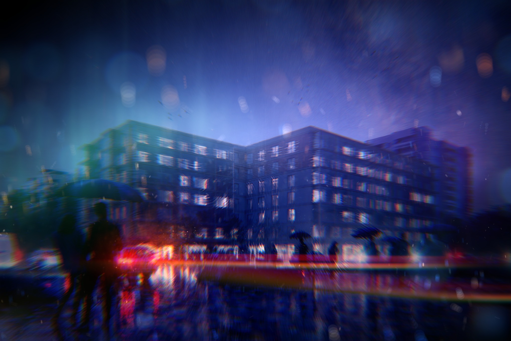 Nocny widok na budynek mieszkalny w deszczu, z rozmytymi sylwetkami ludzi z parasolami i odbiciami świateł w mokrej nawierzchni. Efekt rozmycia dodaje artystycznego charakteru.