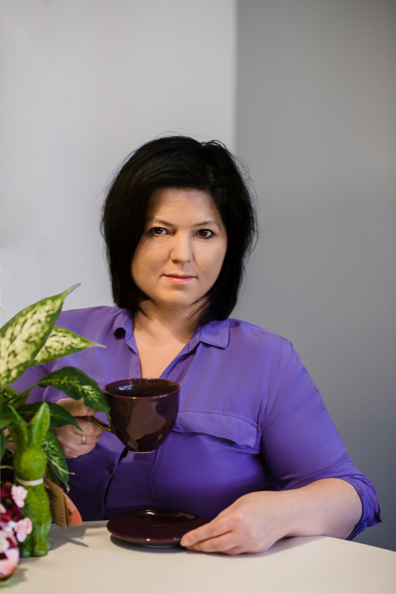 Kobieta w fioletowej koszuli pije kawę z ciemnobrązowego kubka przy białym stole, obok doniczki z zielonym krzewem i dekoracyjnym zajączkiem.