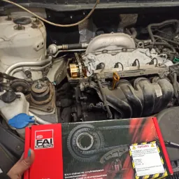 Otwarty silnik samochodowy z widocznym łańcuchem rozrządu i głowicą, obok pudełko z nowym zestawem łańcucha rozrządu FAI Auto Parts trzymane w dłoni z pomalowanym na turkusowo paznokciem.