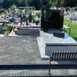 Czarny nagrobek z krzyżem na cmentarzu, otoczony innymi grobami i zielenią. W tle widoczne drzewa. Ujęcie w dziennym świetle.