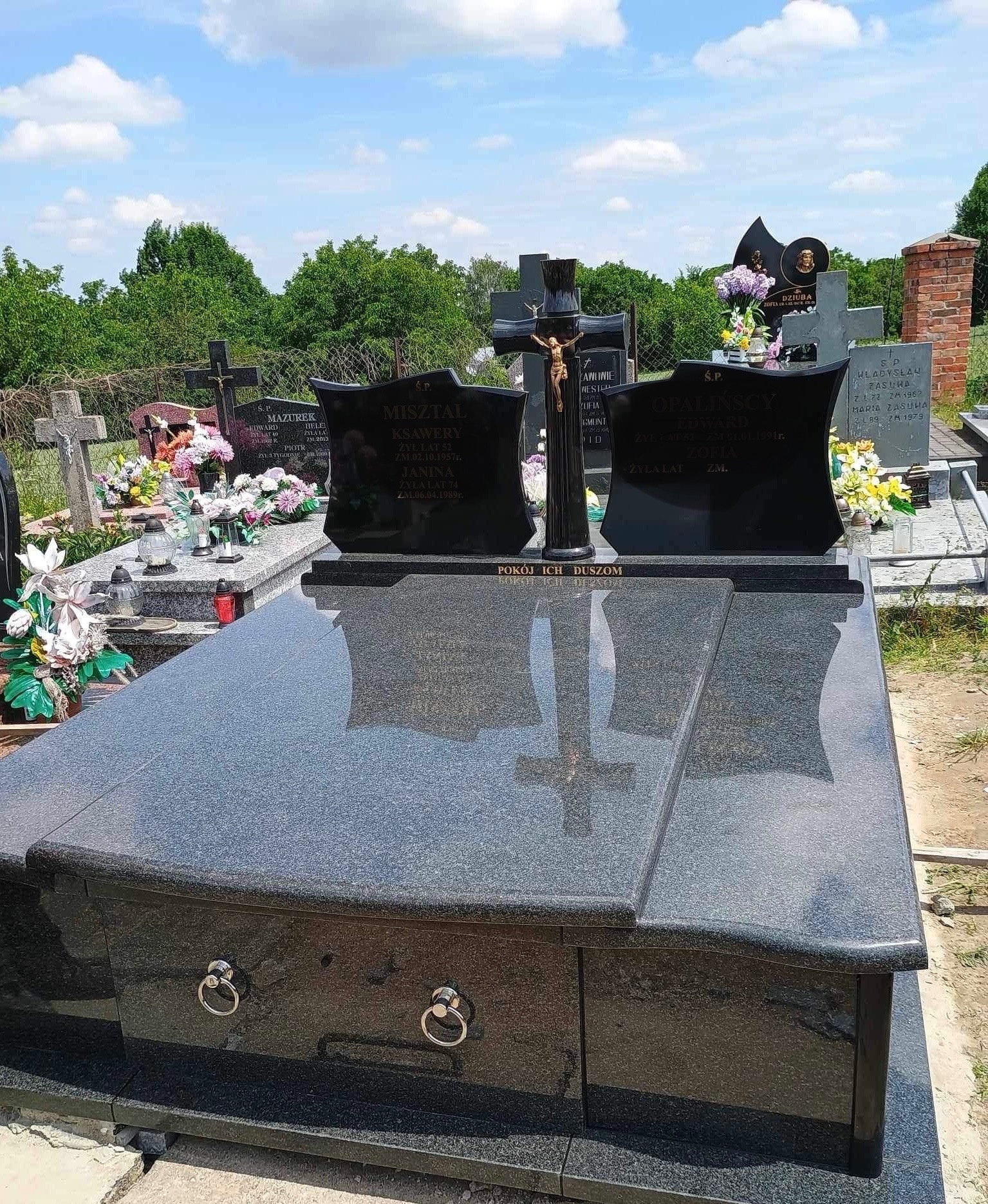 Czarny, granitowy nagrobek z krzyżem w Komarów-Osada, z widocznymi inskrypcjami i metalowymi uchwytami, na tle innych grobów i zieleni cmentarza.