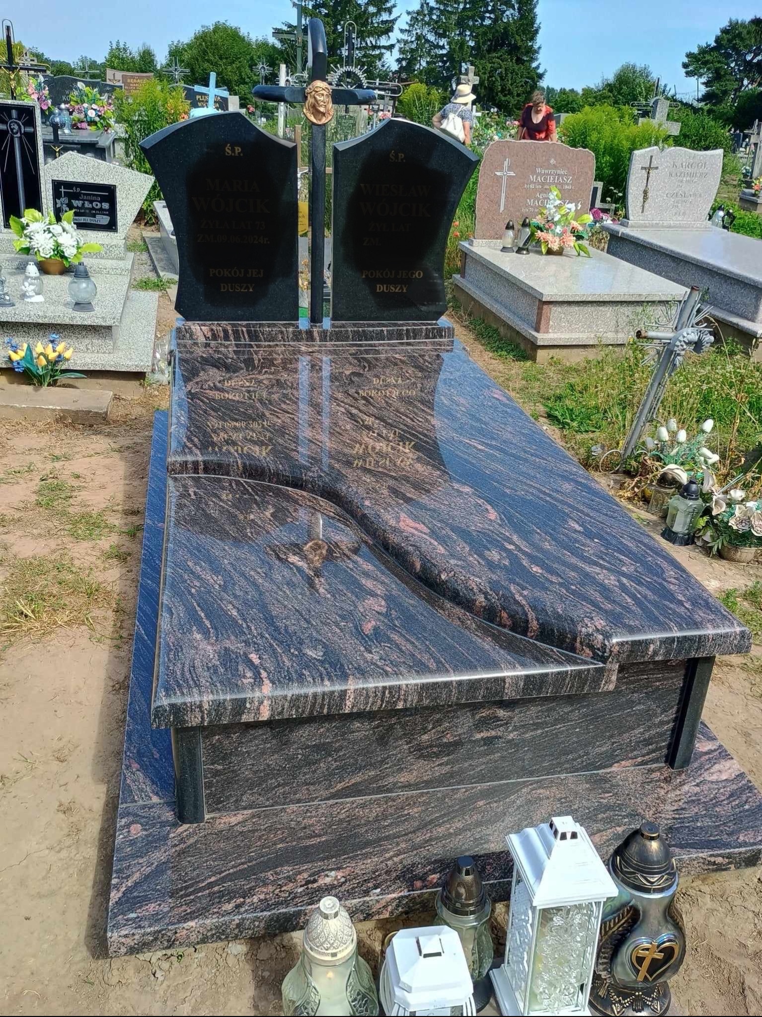 Podwójny nagrobek z ciemnego granitu z czarnymi tablicami inskrypcyjnymi i metalowym krzyżem z wizerunkiem Jezusa, otoczony innymi grobami na cmentarzu.