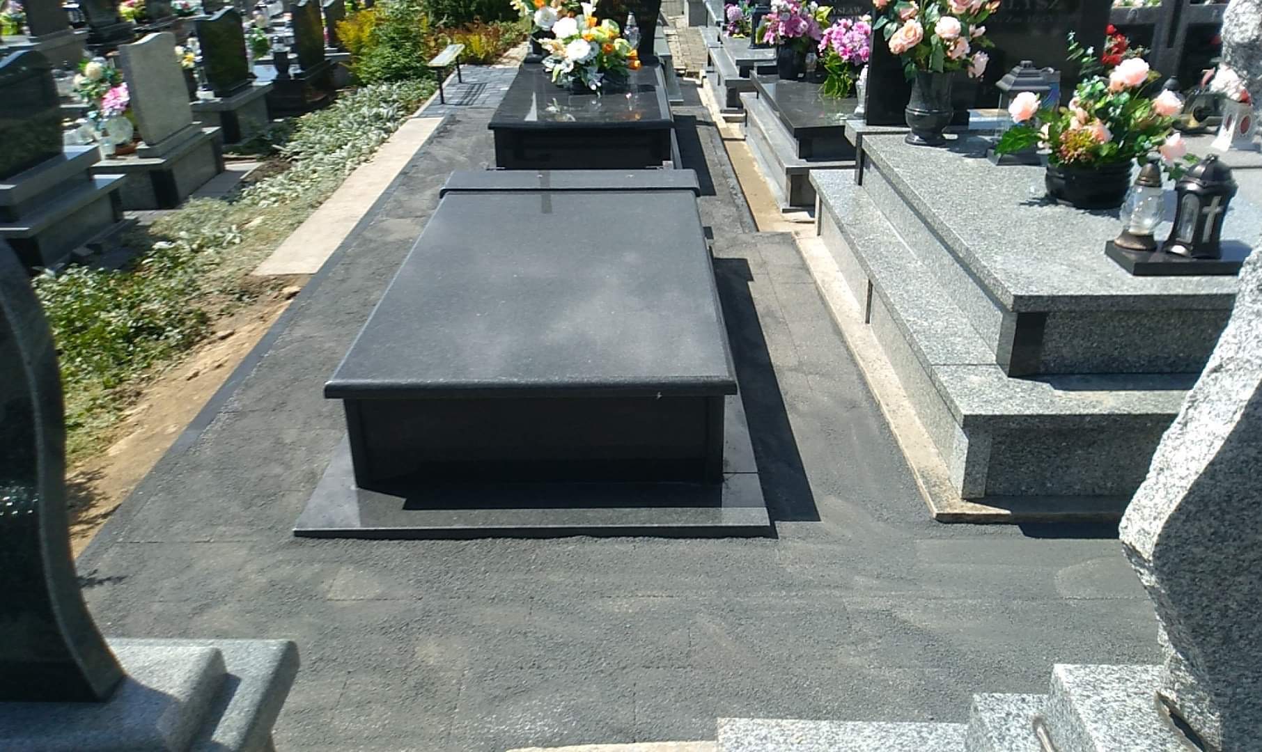 Ciemny, prosty pomnik z płaską płytą nagrobną, otoczony szarą, wyłożoną powierzchnią. W tle inne nagrobki z kwiatami.