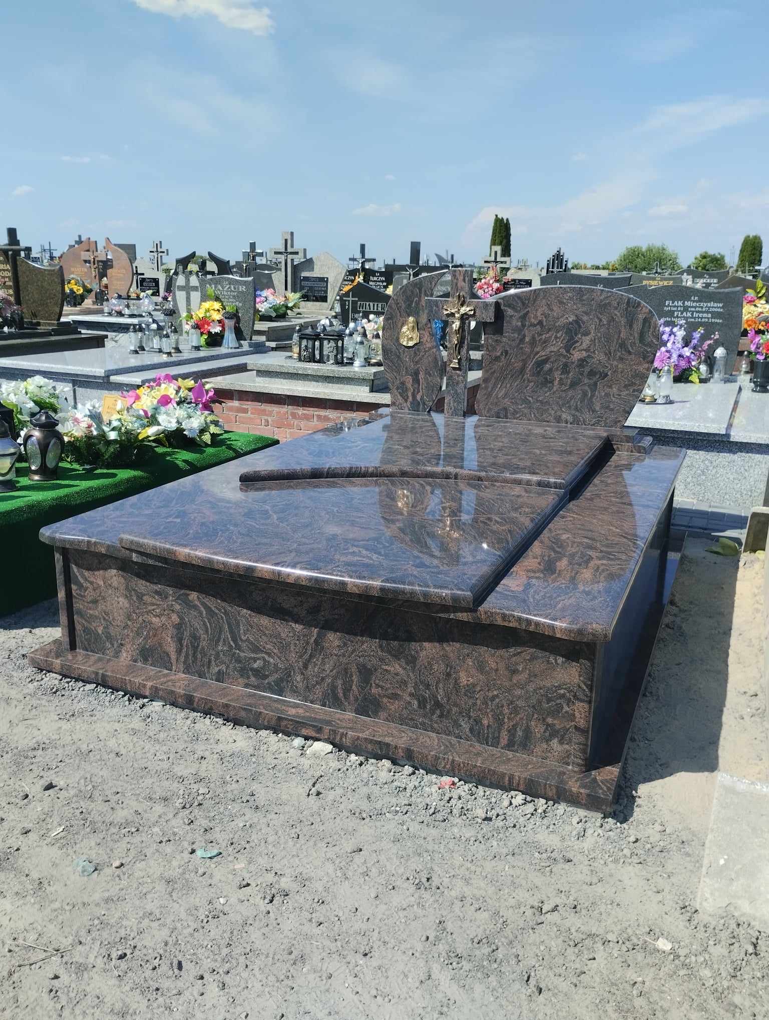Nowoczesny, brązowo-czarny pomnik z polerowanego kamienia na cmentarzu, z krzyżem i złotą ozdobą, na tle nagrobków i błękitnego nieba.