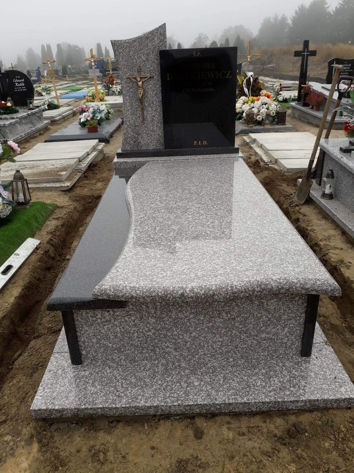 Nowo postawiony, jasnoszary nagrobek z ciemnoszarą tablicą i krzyżem, otoczony świeżo wykopaną ziemią na cmentarzu w mglisty dzień.