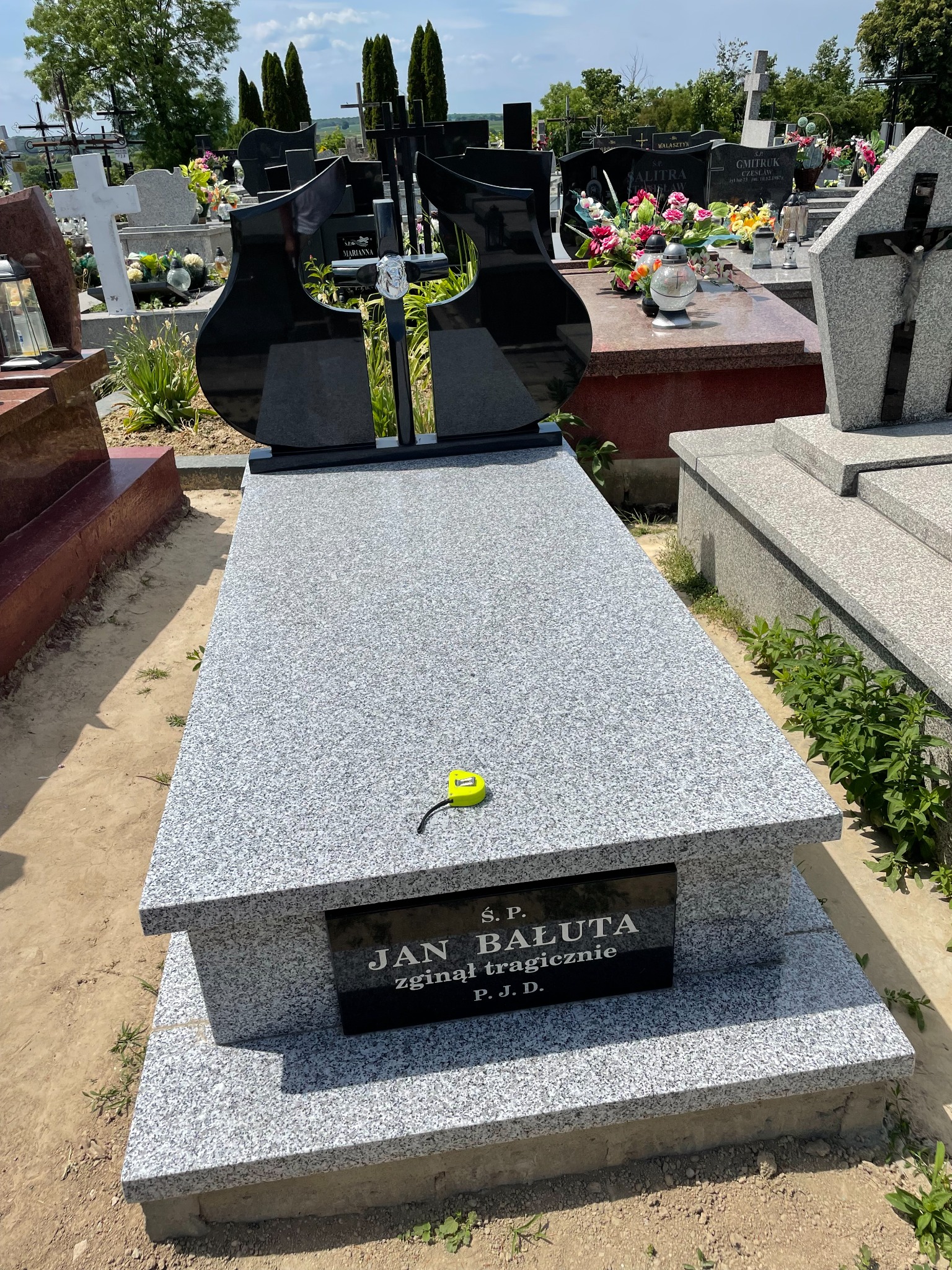 Granitowy nagrobek z czarną tablicą inskrypcyjną 'Jan Bałuta' i nowoczesnym, dzielonym elementem dekoracyjnym w kształcie płomieni, na tle cmentarza z innymi grobami i zielenią, z żółtą miarką...