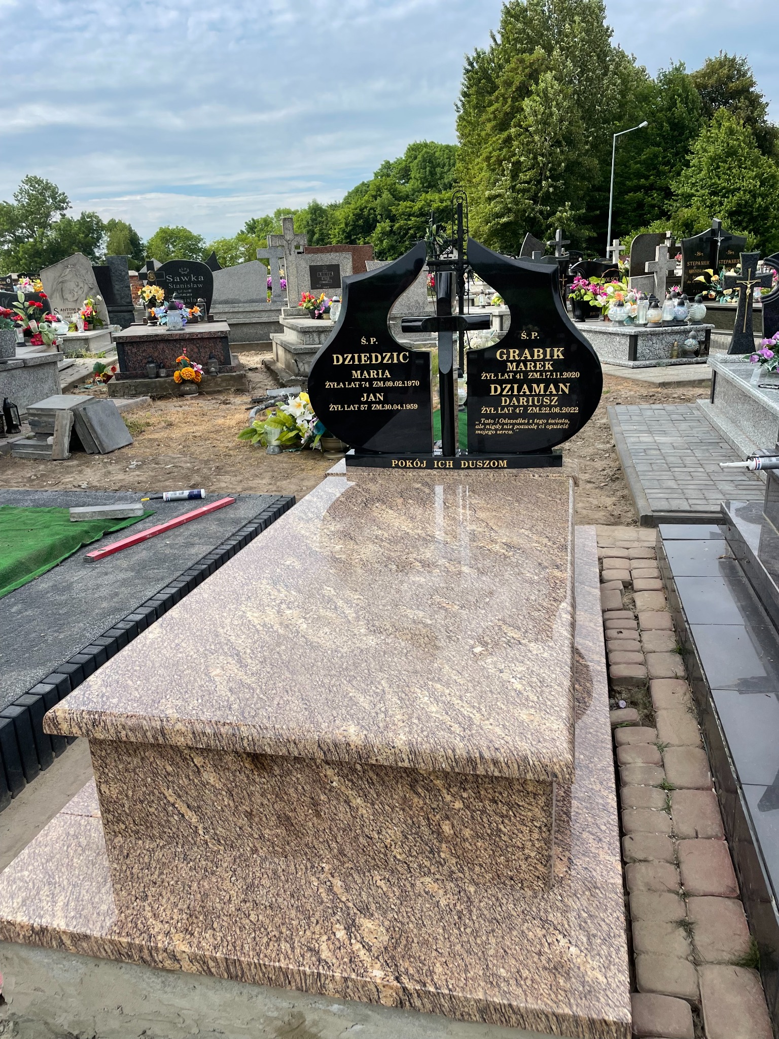 Nowy nagrobek z jasnego kamienia z ciemną tablicą upamiętniającą zmarłych, na tle cmentarza z zielenią i innymi grobami, widoczne narzędzia i materiały budowlane w trakcie montażu.
