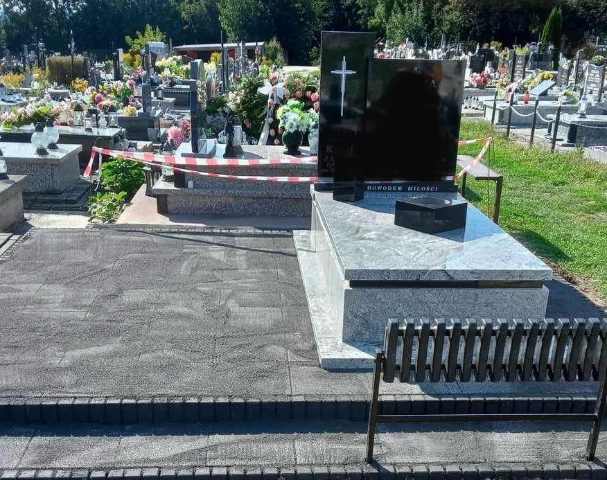 Czarny nagrobek z krzyżem na cmentarzu, otoczony innymi grobami i zielenią. W tle widoczne drzewa. Ujęcie w dziennym świetle.