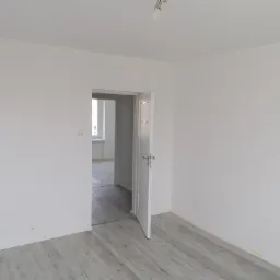 Pusty, świeżo odmalowany pokój z białymi ścianami, otwartymi białymi drzwiami i panelami podłogowymi w odcieniach szarości, widok na sąsiednie pomieszczenie z oknem.