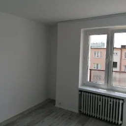 Jasne, puste pomieszczenie po odświeżeniu, z pomalowanymi na biało ścianami i sufitem, szarymi panelami podłogowymi oraz białym grzejnikiem pod oknem z widokiem na budynki mieszkalne.